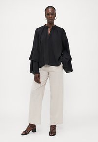Black blouse with ruffled sleeves, V-neck with tie detail. Paired with light beige wide-leg trousers and black strappy sandals. Smooth texture.