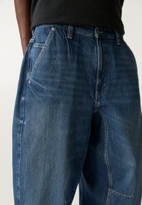 Pantalons en denim bleu avec une coupe ample, des coutures visibles, des poches avant et une finition texturée. Ceinture avec fermeture à bouton.