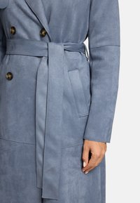 Manteau en daim bleu avec taille ceinturée, design à double boutonnage, grands boutons ronds et poches latérales, présentant une texture lisse et douce.