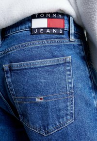 Jeans in denim blu con una toppa posteriore che presenta il logo Tommy Jeans in bianco, rosso e blu. Cuciture visibili e due tasche posteriori.