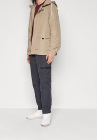 Beige jacka med huva, snörningsdetaljer, två framfickor, bärs över en vinröd skjorta och mörka byxor. Vita sneakers fulländar outfiten.