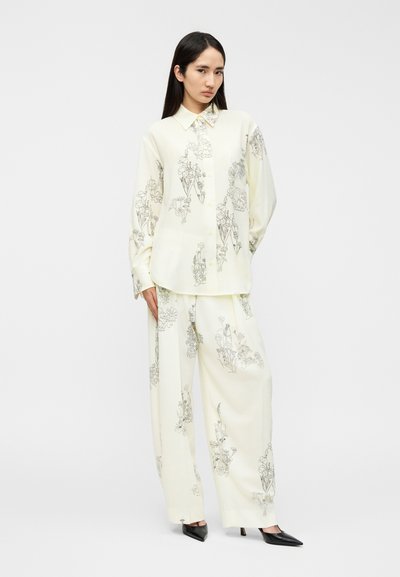 Ανοιχτό κίτρινο, μεταξένιο ταίριασμα πουκάμισου και παντελονιού με φαρδιά μπατζάκια και μαύρα λουλουδάτα σχέδια· πουκάμισο με κουμπιά και γιακά· μαύρες μυτερές γόβες.