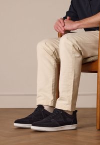 Sneaker in suede blu navy con suole in gomma bianche, caratterizzate da un design a taglio basso e dettagli testurizzati, indossate con pantaloni beige a risvolto mentre si è seduti.