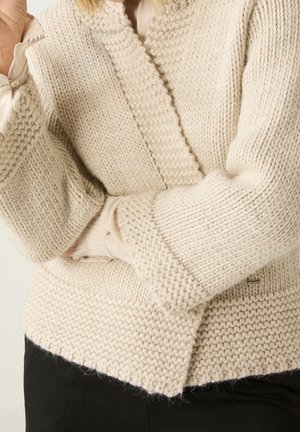 Femme portant un cardigan en grosse maille couleur crème avec des motifs texturés sur un chemisier clair et un pantalon noir, bras croisés.