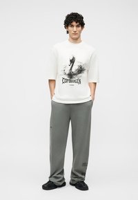 Bărbat tânăr cu păr creț poartă un tricou alb supradimensionat cu grafică de lebădă și textul "Copenhagen", pantaloni largi gri și adidași negri.