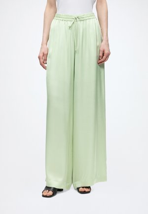 Pantalon large vert clair avec taille élastique et cordon, porté avec des sandales noires à bout ouvert, visible de la taille jusqu'aux pieds.
