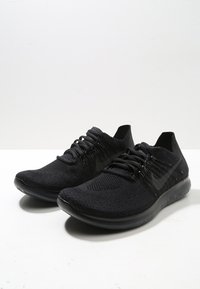 Chaussures de sport légères noires avec une tige en tissu tricoté, des lacets plats et une semelle flexible et texturée. Marque minimale en noir.