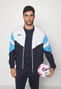 Trainingsjack in wit, marineblauw en lichtblauw, met een ritssluiting aan de voorkant, staande kraag en contrasterende kleurblokken. Model houdt een gemêleerde voetbalsbal vast.
