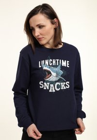 Marineblaues Sweatshirt mit einem Grafikdruck eines Hais und dem Text "MITTAGSSNACKS" in fetten weißen Buchstaben. Weiches Material, Rundhalsausschnitt.