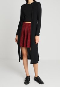 Black ribbed long cardigan layered over a black crop top and a red textured skirt, paired with black flat shoes featuring buckles.