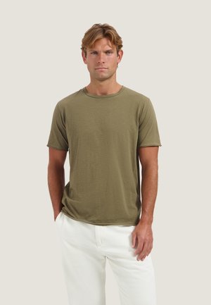 Uomo con capelli castano chiaro, che indossa una maglietta verde oliva a maniche corte e pantaloni bianchi, in piedi con una mano in tasca.