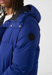 Blauwe pufferjack met een zwarte capuchon, voorzien van een rits aan de voorkant, drukknoopjes en een ronde zwarte logo patch op de linkermouw. Gladde, glanzende stof.