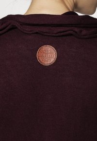 Burgundowy top z teksturowanej tkaniny, z okrągłą skórzaną naszywką z wytłaczanymi detalami przyszytą na tyle przy dekolcie.