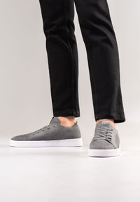 Graue Stoffturnschuhe mit strukturiertem Obermaterial, weißer Gummisohle und sechs Ösen für Schnürsenkel, kombiniert mit schwarzen schmal geschnittenen Hosen.