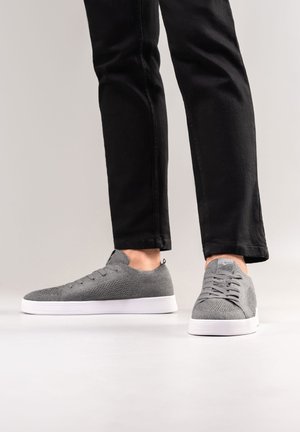 Gray fabric sneakers with a textured surface, white rubber sole, and six eyelets for laces, paired with black fitted pants.