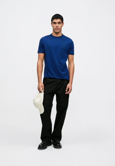 T-shirt bleu marine à manches courtes, coupe droite, associé à un pantalon noir et une casquette de couleur claire, créant un look décontracté.
