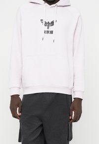 En person som bär en ljusrosa huvtröja med en svart mal-grafik och japansk text, matchad med mörkgrå cargobyxor.