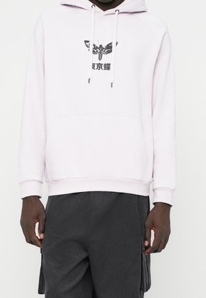 Persona con una sudadera rosa claro que presenta un gráfico de una polilla negra y texto en japonés, combinada con pantalones cargo gris oscuro.
