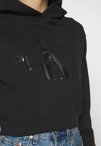 Felpa nera corta realizzata in morbido tessuto, con un logo lucido Calvin Klein sul petto e un design del cappuccio classico.
