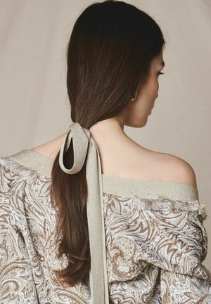 Kvinde med langt mørkt hår bundet tilbage med et beige stofbånd, iført en off-shoulder top med paisley-mønster og frynsekant.