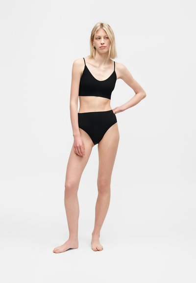 Γυναίκα όρθια ξυπόλητη μπροστά σε λευκό φόντο, φοράει μαύρο κοντό τοπ τύπου καμιζόλας και μαύρο σλιπ ψηλόμεσο, με το ένα χέρι στη μέση.