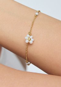 Złota bransoletka łańcuszkowa z kwiatowym wzorem wykonanym z białych koralików, podkreślona złotymi koralikami. Bransoletka spoczywa na skórze w odcieniu ciała.
