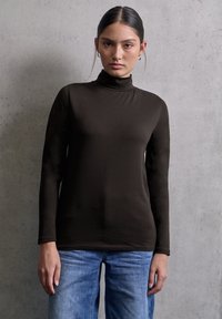 Bruine lange mouwen turtleneck gemaakt van soepel materiaal, met een aansluitende silhouet. Gecombineerd met lichtblauwe jeans, tegen een betonnen achtergrond.