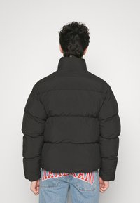 Doudoune noire avec un col haut, présentant un design matelassé et une coupe décontractée. Coutures horizontales larges et notables, portée sur un jean bleu clair.