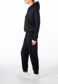 Frau trägt schwarzen Hoodie und passende Jogginghose mit weißen Sneakern, steht im Profil vor weißem Hintergrund.