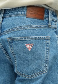 Μπλε τζιν παντελόνια με δερμάτινο ταμπελάκι που απεικονίζει το "GUESS Jeans" στη μέση. Διαθέτει κόκκινο και άσπρο τρίγωνο λογότυπο στην πίσω τσέπη.