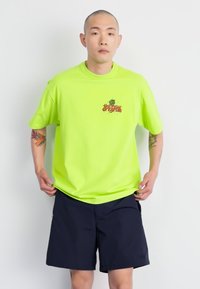 Neon grön bomullströja med "the new originals" grafik på bröstet; matchad med mörkblå shorts med fickor.