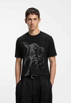 Junger Mann trägt schwarzes T-Shirt mit großem schwarzem Panther-Grafikdruck und dunkle Hose, Hände in den Taschen, vor einfachem Hintergrund.