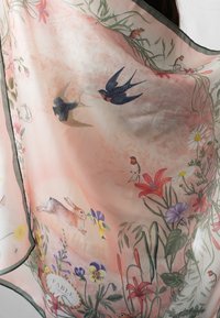 Une écharpe en tulle rose présente des illustrations détaillées d'oiseaux, de fleurs et d'un lapin, avec une bordure vert foncé et une étiquette portant la mention "FABLE".