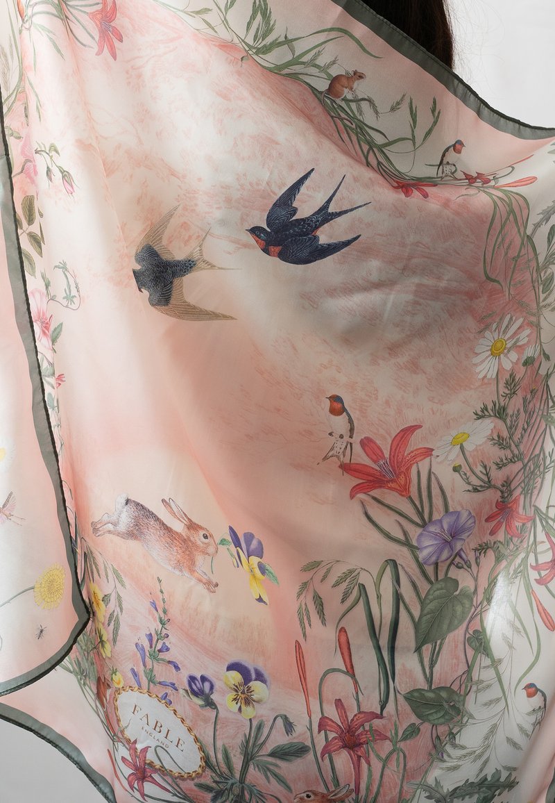 Une écharpe en tulle rose présente des illustrations détaillées d'oiseaux, de fleurs et d'un lapin, avec une bordure vert foncé et une étiquette portant la mention "FABLE".