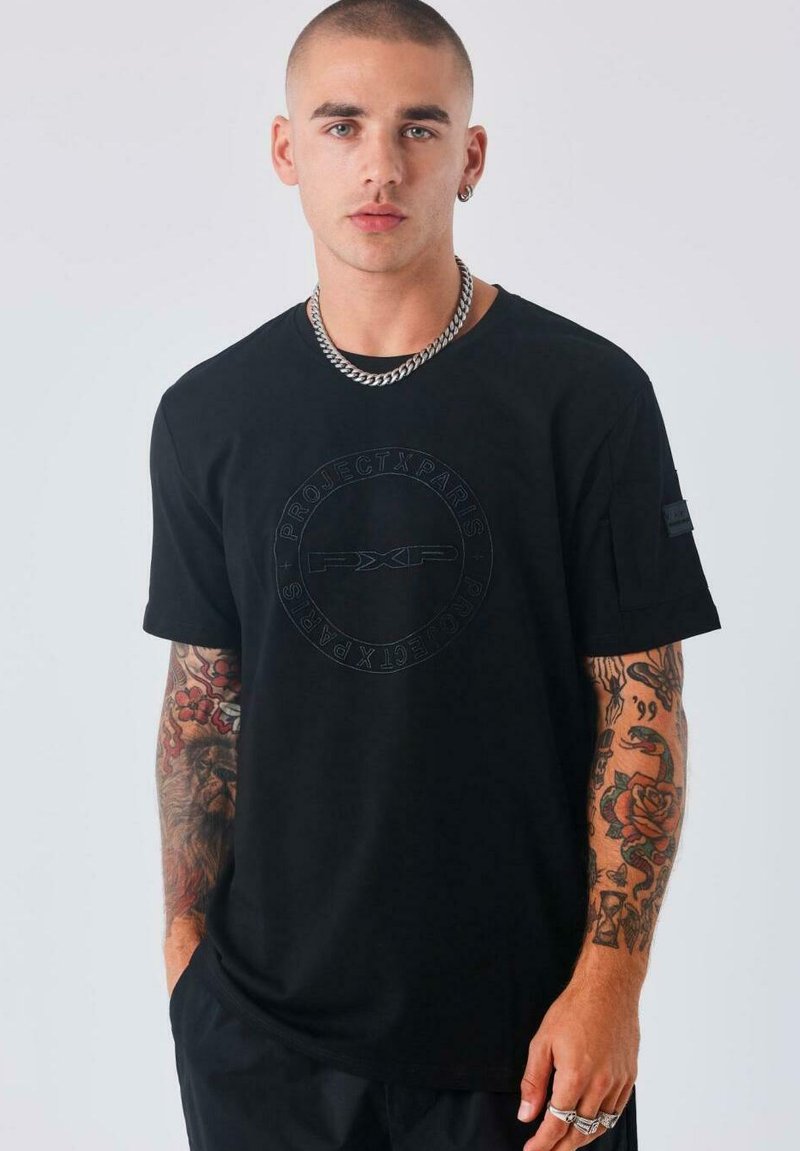 Joven con cabeza rapada que lleva una camiseta negra de Project X Paris, collar de cadena plateada y muestra coloridos tatuajes en ambos antebrazos.