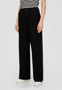 Schwarze weite Hosen mit Knopfverschluss, Gürtelschlaufen und Seitentaschen, kombiniert mit weißen Turnschuhen für einen lässigen Look.