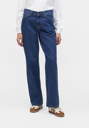 Persona che indossa jeans blu a gamba dritta, camicia bianca a maniche lunghe e sneakers marroni e bianche, in piedi su sfondo bianco.
