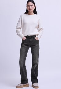 Suéter de canalé crema, jeans rectos grises y zapatos de plataforma beige. Modelo de pie con las manos en los bolsillos, fondo neutro.