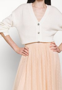 Cardigan bianco a maglia con scollo a V e tre bottoni, abbinato a una gonna in tulle pesca con piccoli motivi a stelle bianche e una texture fluida.