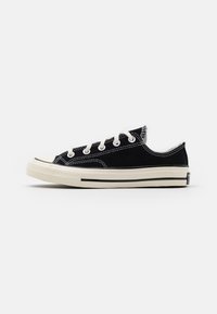Sneaker nera in canvas bassa con lacci bianchi, suola in gomma bianca e striscia nera a contrasto, vista di lato su uno sfondo bianco.