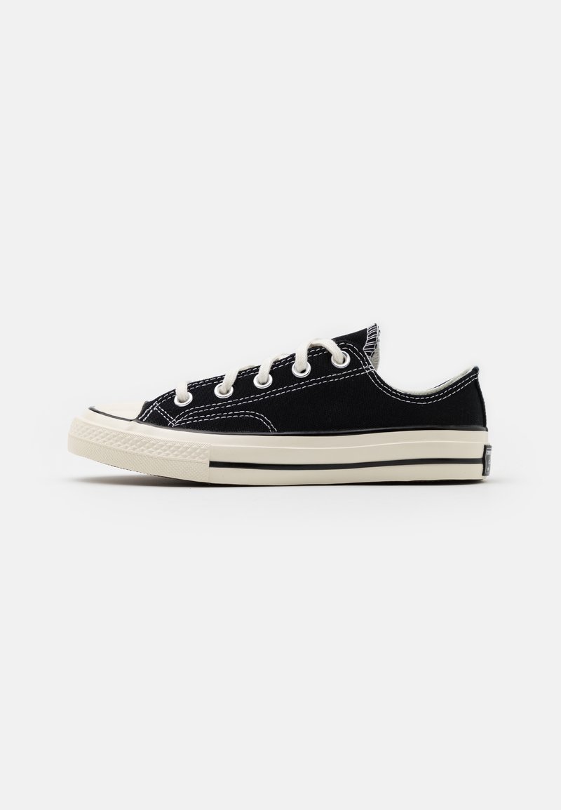 Sneaker nera in canvas bassa con lacci bianchi, suola in gomma bianca e striscia nera a contrasto, vista di lato su uno sfondo bianco.