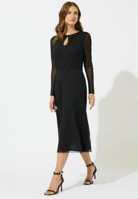 Abito midi nero con maniche lunghe trasparenti, presenta un collo a chiave e un motivo a pois metallici. Abbinato a tacchi a spillo neri con cinghie.