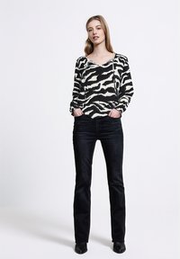 Blusa a righe zebra bianco e nero con maniche lunghe, abbinata a jeans blu scuro e stivali neri alla caviglia, su uno sfondo chiaro.