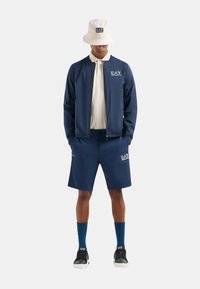 Completo sportivo navy composto da un bomber con zip, pantaloni corti abbinati, un cappello a secchiello beige chiaro, calze blu e sneakers nere con suola bianca.