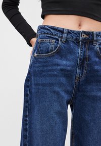 Modré denimové džínsy s voľným strihom, vysokým pásom, dvomi prednými vreckami a nenápadným kontrastným prešívaním. Bez viditeľného opotrebovania.