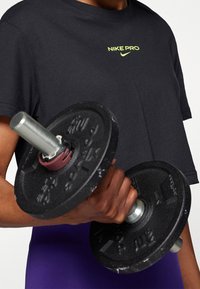 Sort Nike Pro t-shirt med et neongrønt logo, båret af en person, der holder en sort vægtplade med et sølvhåndtag og pink detaljer.