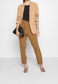 Blazer beige de un botón, top negro ajustado, pantalones marrones de cintura alta con lazo en la cintura, y clutch negro. Chanclas negras.