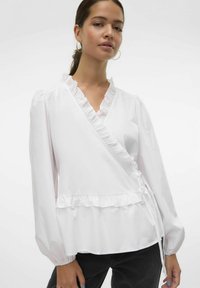 Camicia bianca con scollo e orlo arricciati, maniche lunghe a sbuffo e dettaglio con lacci laterali, realizzata in tessuto morbido, abbinata a pantaloni scuri.