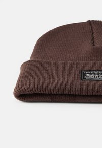 Gorro de malha canelada castanho com uma borda dobrada e um rótulo retangular preto com texto branco e designs gráficos. Textura suave.