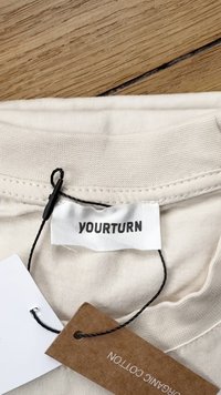 Etiqueta de vestuário de algodão bege com a marca "YOURTURN". Costura preta anexada a uma etiqueta castanha com a inscrição "ALGODÃO ORGÂNICO". Textura de tecido suave.